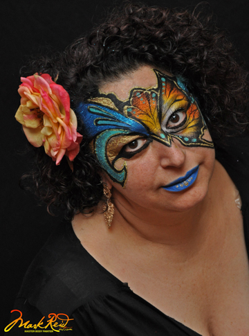 woman with an intricate mask that has been painted on her in bright colors with a flower behind her ear